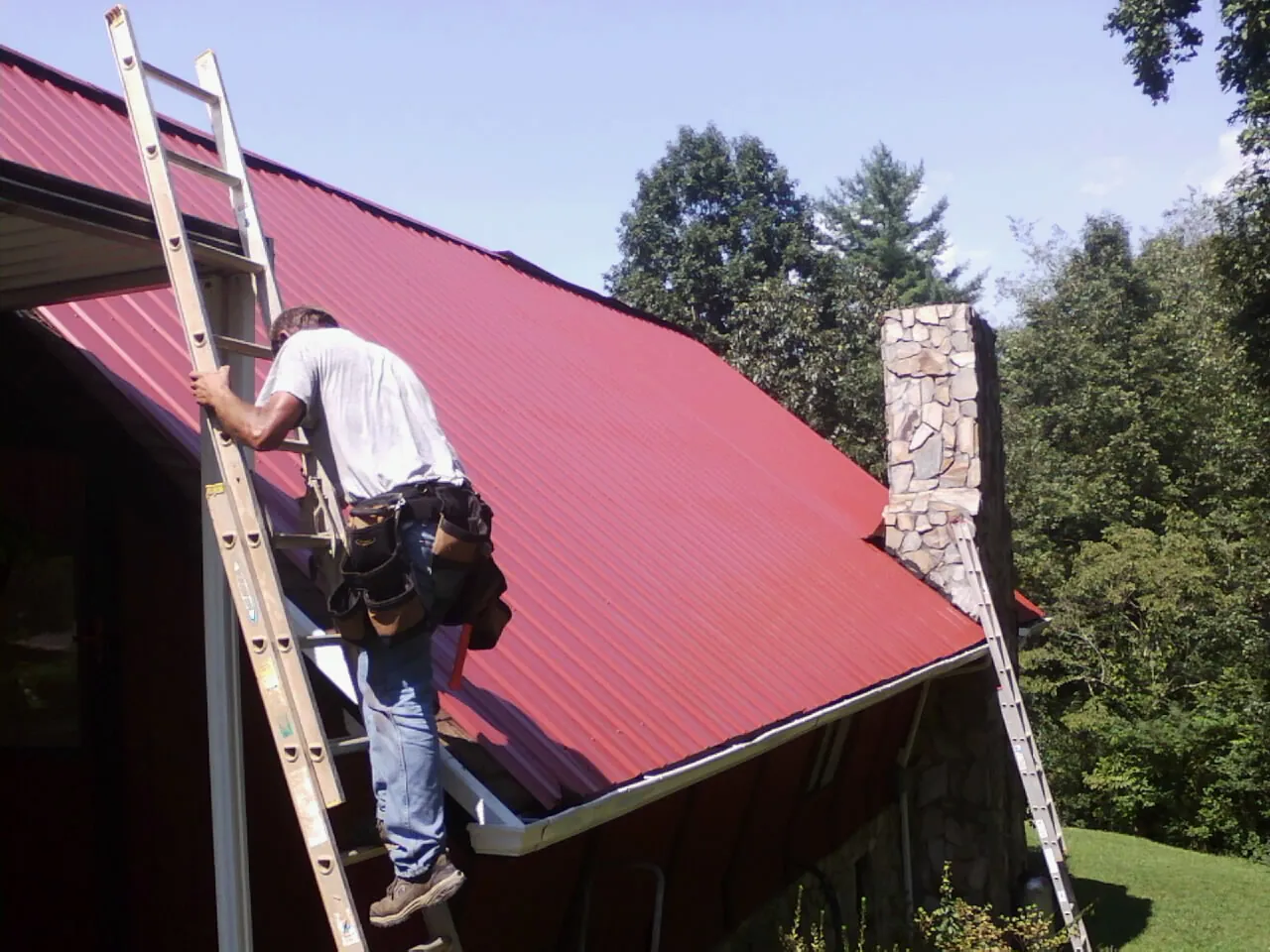 Roofer on a ladder beside a red metal roof and stone chimney — Soffit & Fascia Repair by Roofing Brothers in Egypt